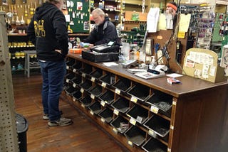 An employee standing behind and old wooden counter with mental bins. A customer on the other side completes a purchase. Walls in the background are covered with paitn brushes, stains, and wrneches.
