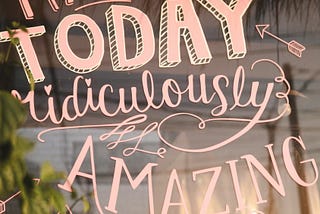 Motivational hand-lettered sign in pink, from a café window that reads “Make Today Ridiculously Amazing,” with playful arrows and swirls, displayed on a reflective glass surface with plants and soft light in the background.