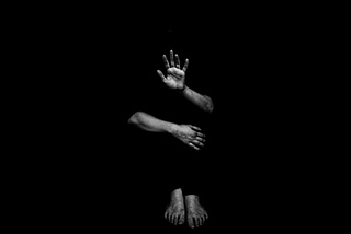 man in darkness with hand held up and arm wrapped around legs