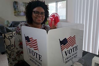 A picture of me, Aria, sitting at a table with a voting booth that says “Vote from home booth 2020.”