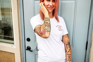 Tall, heavily tattooed caucasian woman with pink hair in a white tee shirt standing outside before an eggshell blue door.
