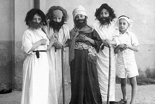 Five boys dressed in biblical costumes standing in a row outside in front of a blank wall. The ground is covered in dirt. Four are wearing wigs and fake beards. Two hold canes. The boy in the center stands out, dressed in a patterned, dark full-length garment, while the others are dressed in solid, white full-length garments. One boy on the far right is wearing white shorts and sandals.