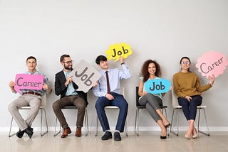 Left to right, three men and two women sit on chairs holding signs saying “career” or “job” presented in coloured speech bubbles.