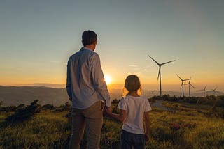 A father and his young daughter watching the sunset over a rural landscape that includes modern day windmills in the background.