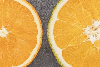 Five cross sections of citrus fruits arranged in ascending order of diameter on a textured grey background. From left to right, they appear to be a lime, a lemon, and orange, a larger orange citrus fruit, and a red grapefruit.