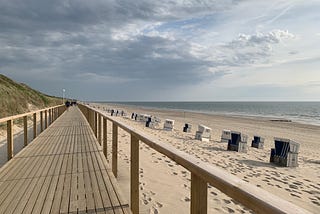 A long wooden boardwalk with wooden rails runs along the beach, with basket beach chairs on the sand, and the dark sea beyond.