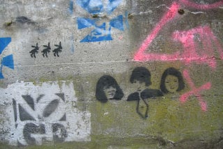 A photo by Moira Cloney, showing green, mouldy concrete covered with graffiti, including a stencil of Sleater-Kinney’s faces.