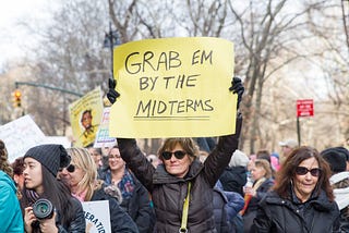 A white woman holds a sign at a peaceful protest that reads “Grab them by the midterms”