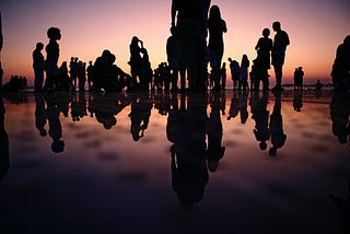 Group of people gathered at a beach at dusk, their silhouettes reflecting in the water.
