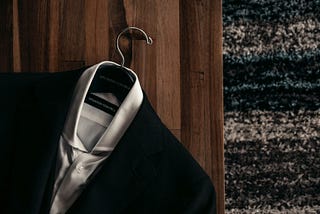 Black suit jacket over white shirt on a hanger against a brown door with black, gray, and white carpet on the right. The tag on the shirt and hanger says Situation Control.