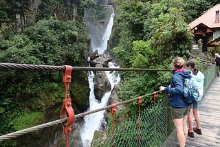 Two women stand on a cable pedestrian suspension bridge gazing at a large waterfall surrounded by lush greenery in the mountain forests of Ecuador. They are wearing hiking shoes, shorts, rain jackets, and backpacks. Their backs are to the camera.