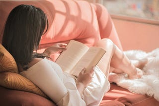 girl with dark hair wearing white shirt reading a book and sitting on pink sofa