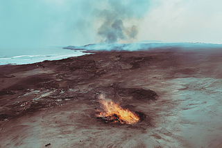 A small fire smoulders on a piece of arid land, with the sea in the distance, and smoke and ash floating in the air.