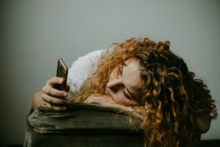 A woman with long curly red hair is slumped over a cushion, looking at her mobile phone. She seems sad or melancholy as she stares at her phone.