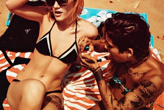 A blonde girl in a black bikini and a brunette girl with tattoos sit on towels and smoke marijuana on a beach.