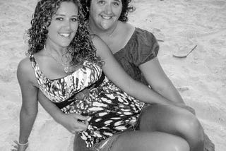 Black and white photo of the author, a young woman, sitting across her mothers lap on a sandy beach.