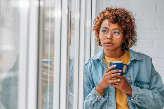 Woman holding a coffee cup looking wistfully out the window.