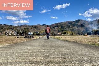 A man is turning his back at the camera, walking in the middle of a deserted road in the Japanese countryside, towards the mountains. The sky is blue.