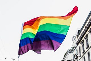 Pride flag waving in front of a city building. Image is black and white, except for the rainbow colors of the Pride flag.