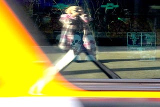 A blonde woman in a pink, brown and white plaid mid-length coat taking a photo of herself in a passing bus window.