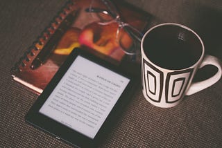 Photo of an e-reader, notebook and coffee cup on wooden surface