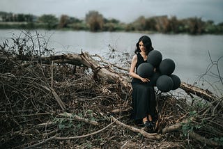 Woman sitting on a tree wearing a black dress and holding black balloons