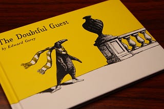 Cover of “The Doubtful Guest” by Edward Gorey (1957) on wooden desk. Cover shows a black penguin-like creature wearing a scarf and sneakers, standing alone on a porch beside an urn.