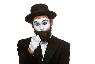 Photo of a mime artist/jester pointing a white-gloved finger at the camera. He has a black beard and wears a hat.