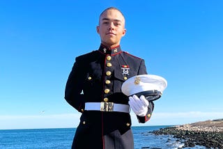Young man wearing the Dress Blues uniform of the US Marine Corps, formal white hat balanced on his forearm. Behind him is the ocean shore and a clear blue sky.