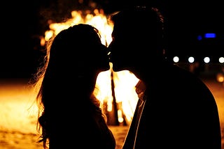 A sexy teenage girl and guy share a secret romantic kiss silhouetted in front of a bonfire on a riverside beach on a hot Summer night.