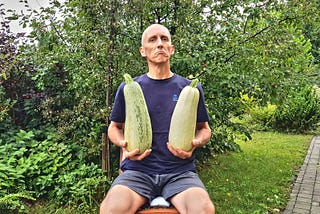 The author, József Manhertz, sits on a chair in his garden in summer in green shorts and a dark blue T-shirt. Lush green bushes and trees surround it. He is holding two huge zucchini in both hands. Meanwhile, he looks at the camera with his head held high, with a funny forced proud expression.