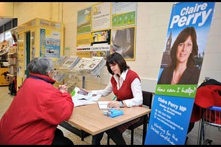 Former MP Claire Perry conducting a constituency surgery meeting with a constituent in a public building with a poster to the left which says “Claire Perry (headshot) How can I help?”