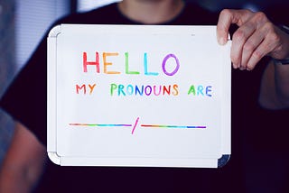 A light-skinned human being in a black shirt holds up a sign with rainbow text reading “HELLO MY PRONOUNS ARE”