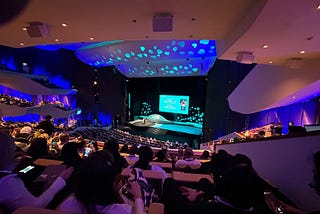 There is a stage in the center of the image and sitting stands around it. There is a screen on stage and people sitting on the the stands.