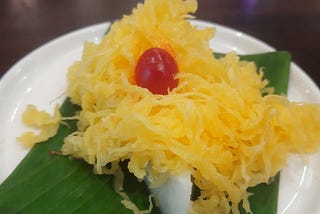 On a white plate, over a banana leaf yellow threads are placed on a custard-texture pudding with a cherry on the top.