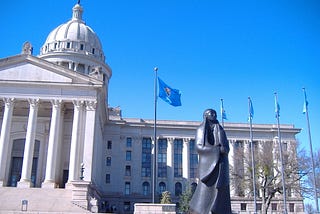 Bronze sculpture of a Native American woman in front of the neoclassical facade of the Oklahoma State Capitol building.