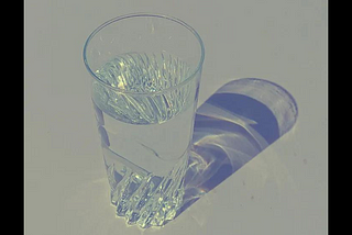 A black-bordered photograph of a glass of water, which casts a hazy, dusky blue shadow onto a light grey surface.
