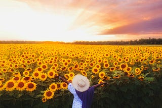 A woman in a white dress raises her arms in a field of sunflowers at sunset