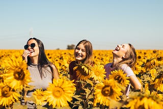 an image of three women smiling and laughing in a sunflower field.