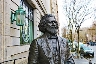 Bronze statue of a man from the nineteenth century. He’s looking forward, and stands next to a decorative copper lamp.