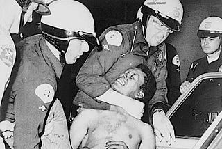 A Black man is shown being held in a headlock by a white policeman as other white policeman look on.