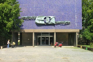 Front face of a concrete building, purple in colour, with a stylized transistor sculpture above the entrance.