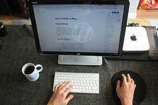 A person working on a blog post about using Gutenberg and Block Editor in WordPress on a desktop computer. The setup includes a keyboard, mouse on a mouse pad, a coffee mug, and a Mac Mini on the desk.