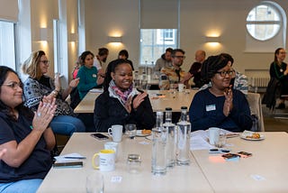 Network members seated at tables in a bright conference room, clapping and smiling during a DLN in-person meetup. The group includes diverse individuals with cups, water bottles and notepads on the tables. Large windows and soft lighting create a welcoming atmosphere.