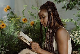 A Black woman with box braids reads a book, behind her are yellow flowers.