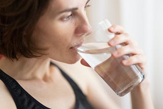 A young woman with short brown hair and wearing a black tank top is drinking a glass of water.