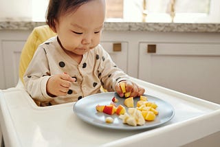 Photo of a baby in a high chair with a plate of cut up foods in front of her.