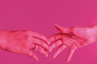 Two hands reaching for one another, seen under pink light against a bright pink background