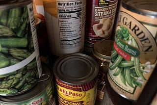 A close up of random stacked cans, like green beans, soup, and asparagus. Not one can of black olives is seen.