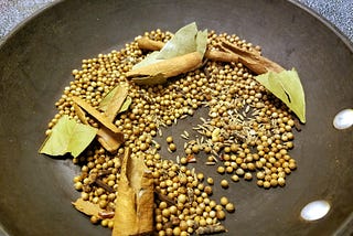 A variety of whole spices, such as bay leaves, cinnamon sticks, coriander seeds, and cumin seeds, is spread out on a black pan. The mixture suggests a preparation for roasting or toasting the spices to enhance their flavors before use in a recipe.